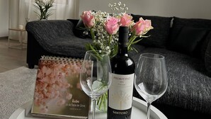 Un salon avec une table ronde blanche, une bouteille de vin, deux verres à vin, un arrangement floral, un livre, une horloge et une lampe.