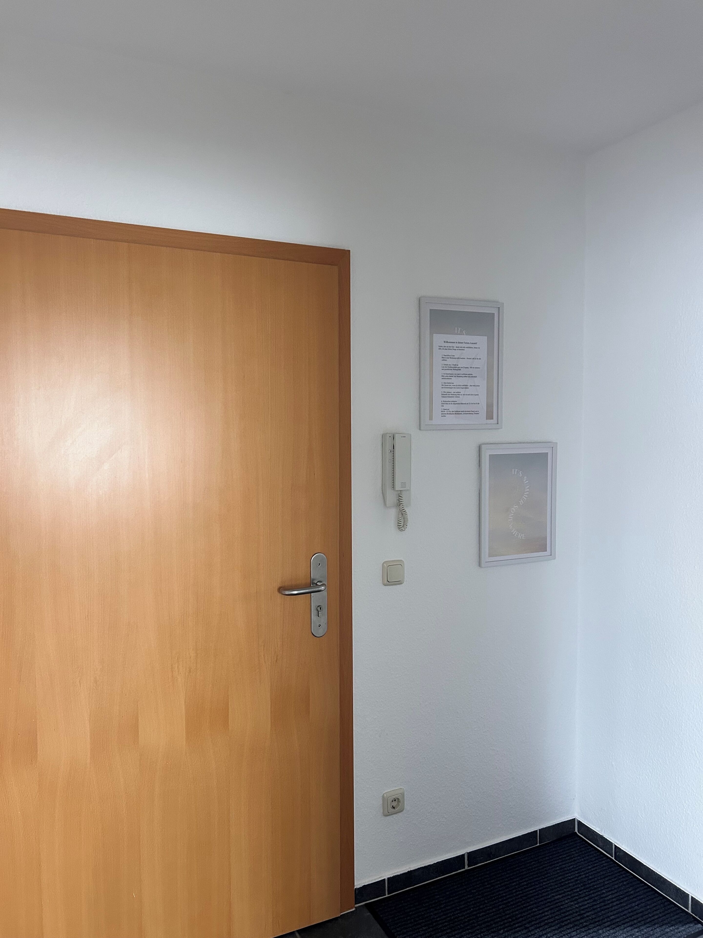 Eine Holztür mit silbernem Griff, eine weiße Wand mit zwei gerahmten Schildern, ein Telefon und ein Lichtschalter.