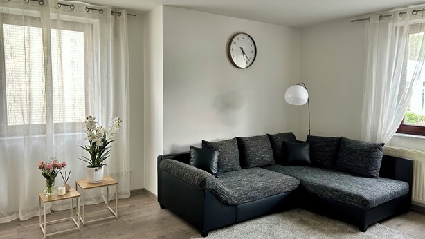 Un salon moderne avec un canapé d’angle gris foncé, une horloge murale ronde, un lampadaire et une petite table sur laquelle est posé un vase de fleurs.