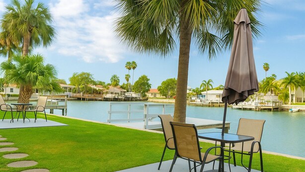 Zona junto a la piscina con mobiliario exterior, una palmera y vistas al agua y edificios en el fondo.