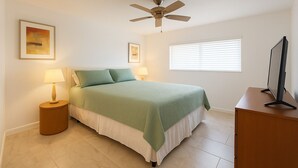 Un dormitorio con una cama, dos mesitas de noche con lámparas, un ventilador de techo, una ventana con persianas y un televisor sobre un mueble.