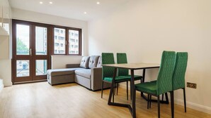 A modern living room with a grey sectional sofa, a wooden dining table with four green chairs, and a large window with a view of buildings. - Spectacular flat in the heart of Shoreditch (London)