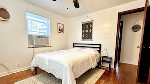 A bedroom with a bed, a ceiling fan, a window with blinds, a wall-mounted air conditioner, a rug, and a door. - Charming 2-bedroom on the West Side! Pet-friendly and quiet! (Cincinnati)