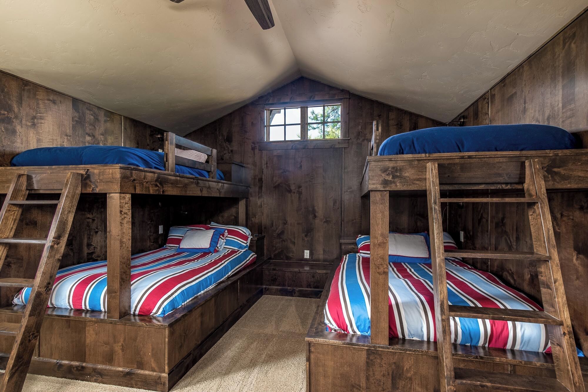A rustic room with two bunk beds, wooden walls, a small window, and a ceiling fan.