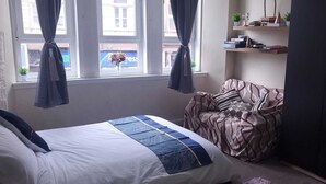 Un dormitorio con cama, sofá, ventana con cortinas y una estantería con libros y objetos decorativos.