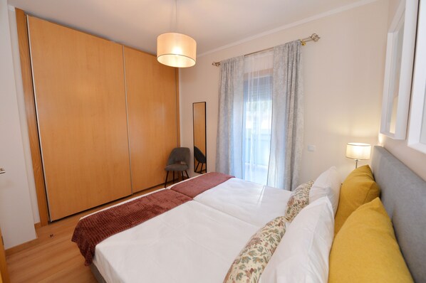 Un dormitorio con cama, una silla, un armario y una ventana con cortinas.