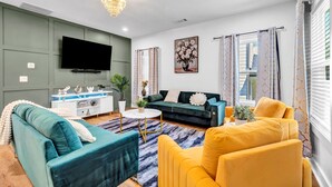 Un salón con un sofá azul verdoso, un sillón amarillo, un sofá verde, una mesa de centro blanca y un televisor de pantalla plana montado en la pared.