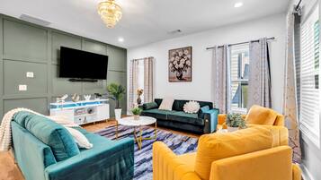 A living room with a teal sofa, a yellow armchair, a green sofa, a white coffee table, and a flat-screen TV mounted on the wall.