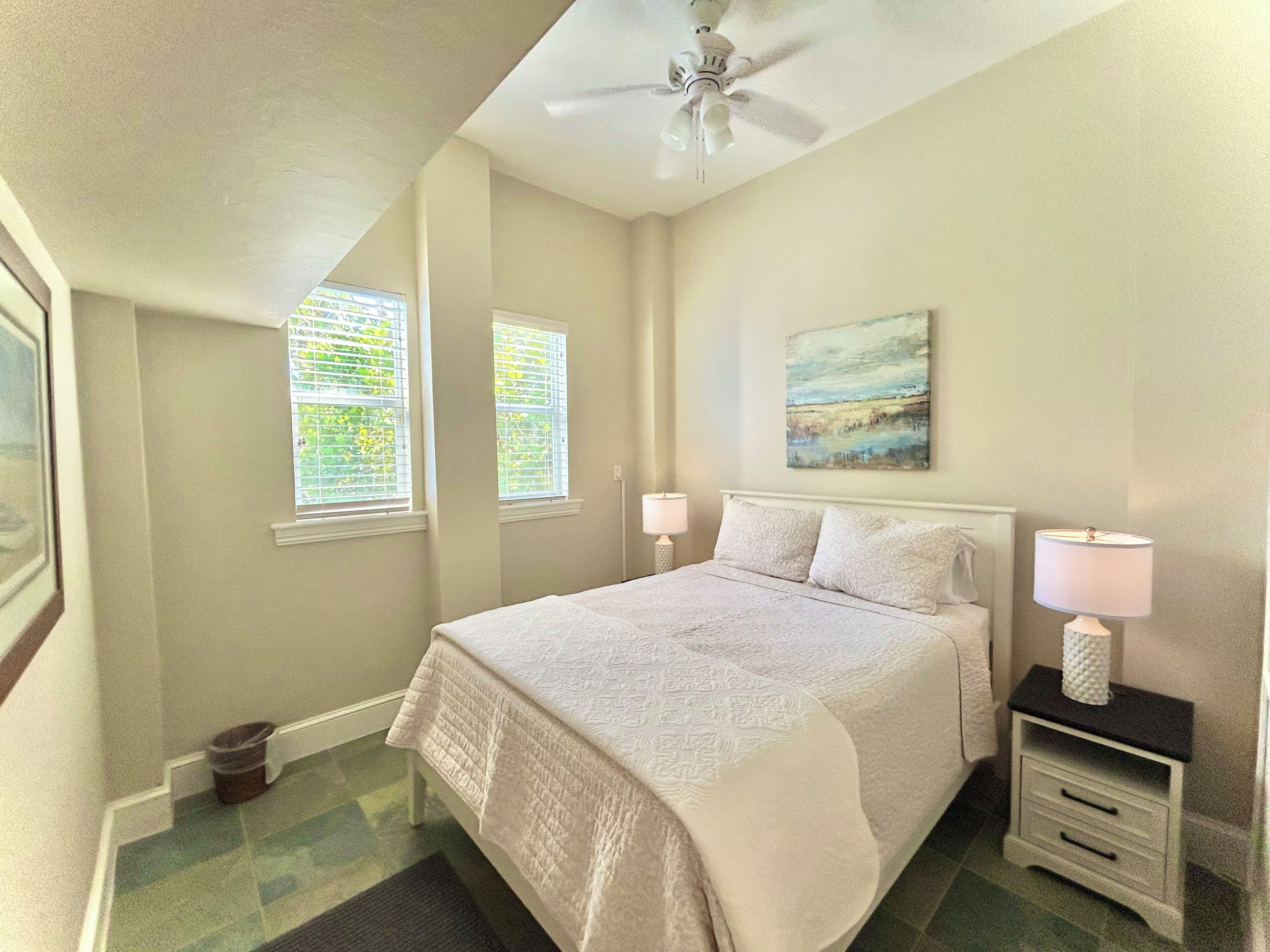 A bedroom with a bed, two bedside lamps, a ceiling fan, and a window with blinds.