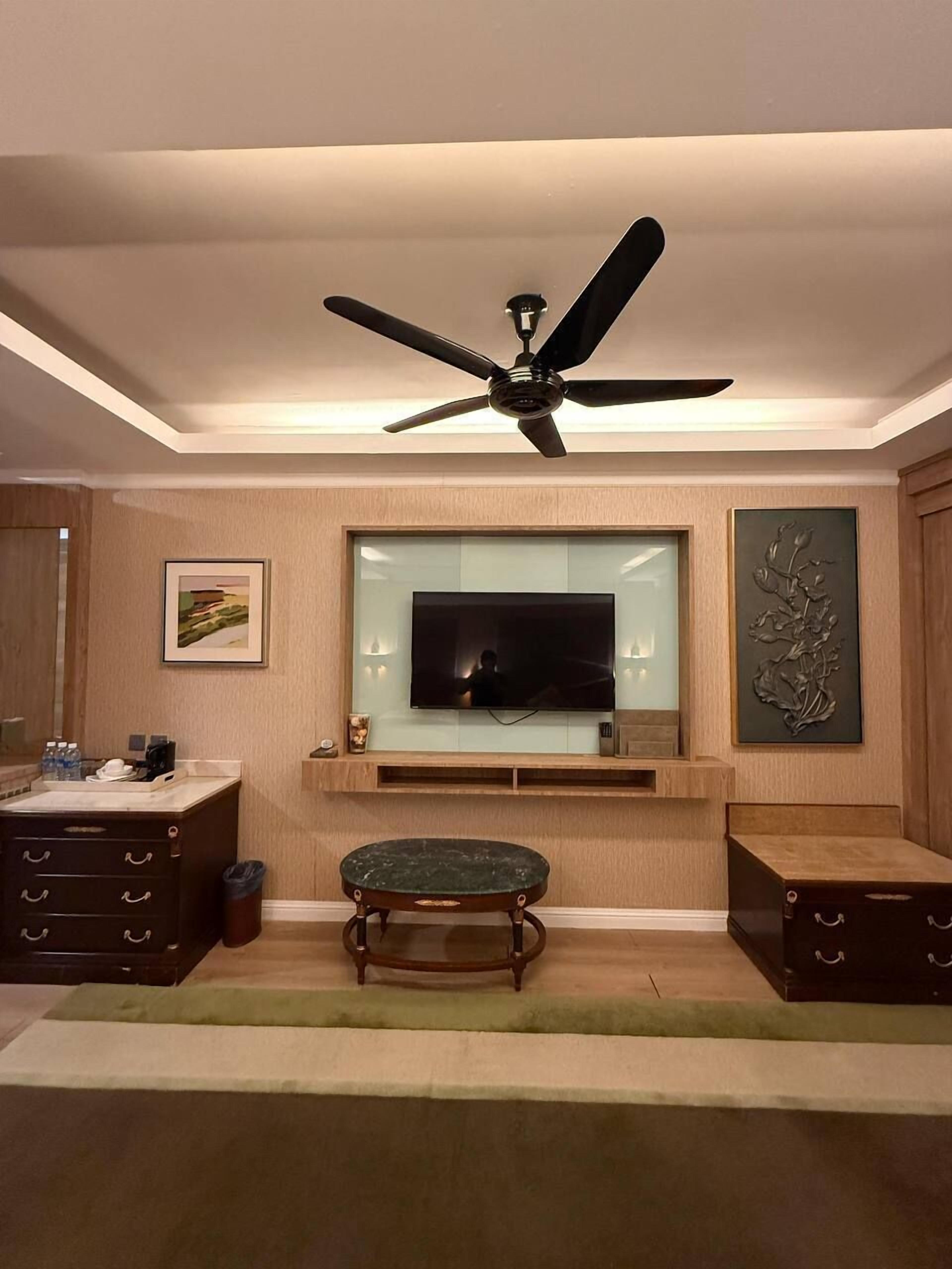 A modern living room with a ceiling fan, a flat-screen TV mounted on a wooden wall, a small round table, and a cabinet with drawers.