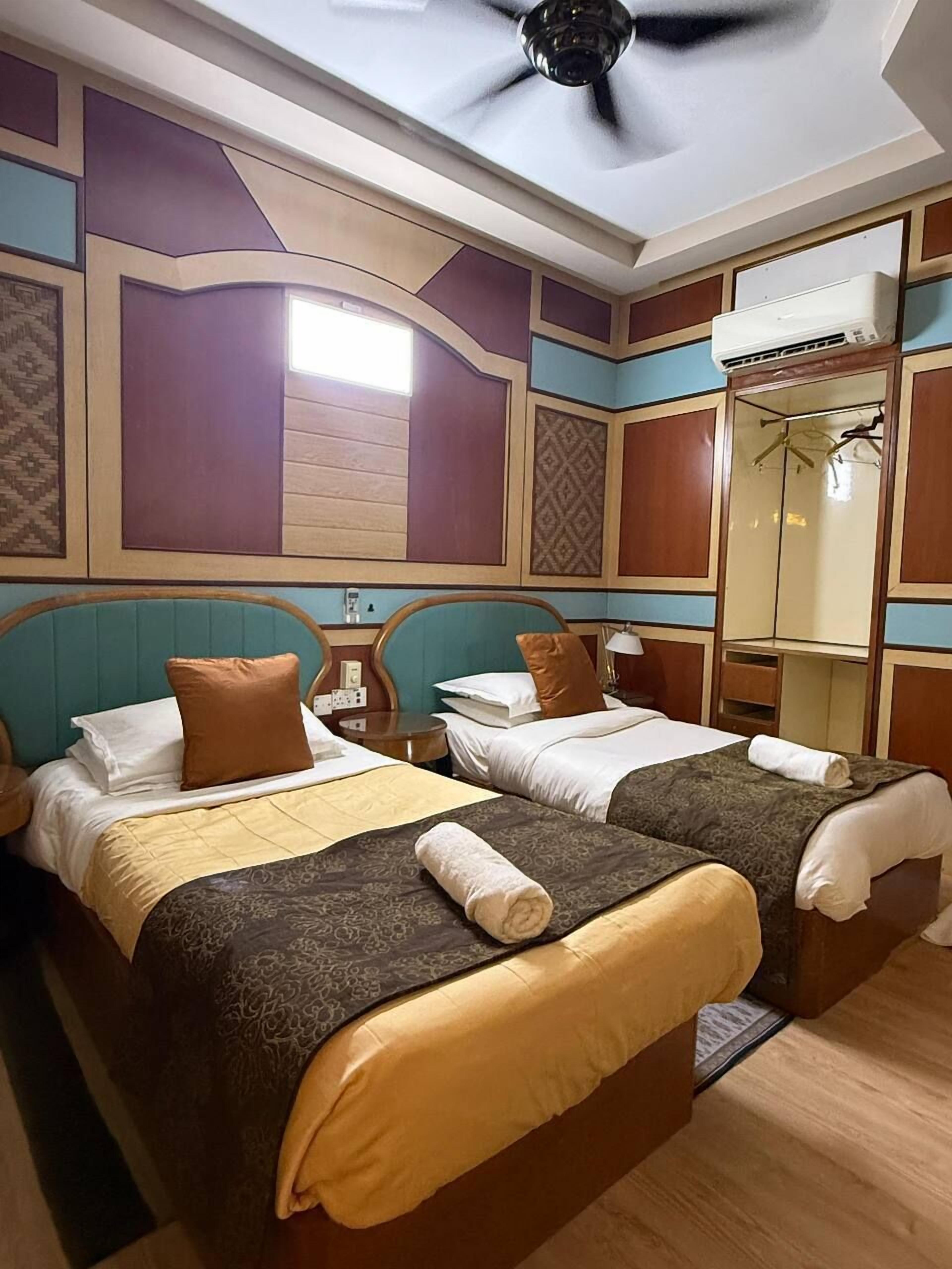 A hotel room with two beds, a ceiling fan, a wardrobe, and an air conditioning unit.