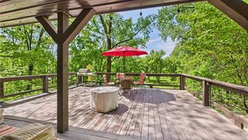 A wooden deck with a round table and chairs, a red umbrella, and a wooden bench.
