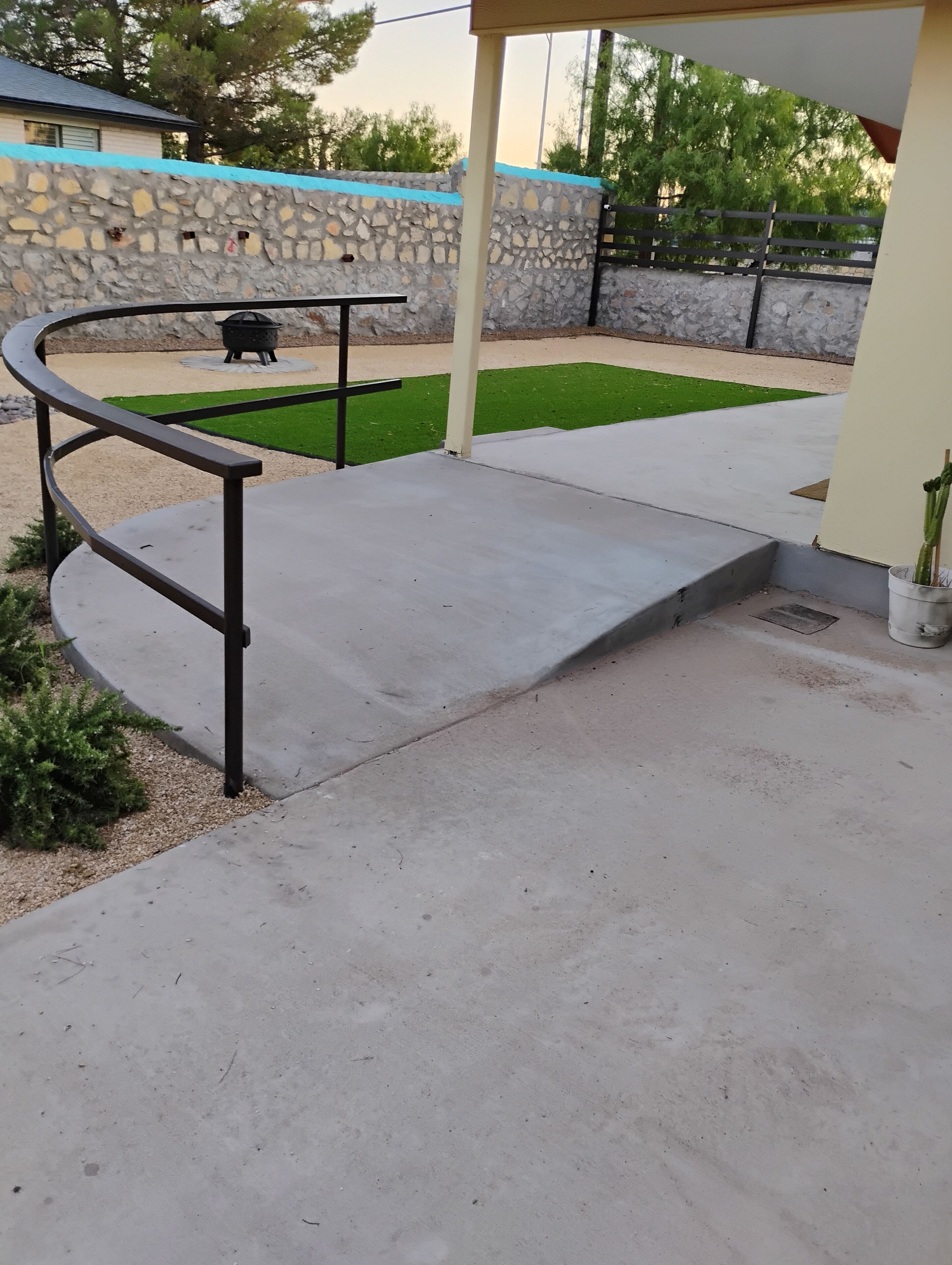 Un patio de concreto con barandilla de metal, pared de piedra y un pequeño césped verde.