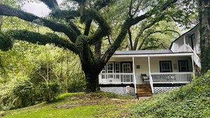 A white house with a porch, surrounded by lush greenery and a large tree. - Cici's Guest House Top rated stay in Laurel (Laurel)