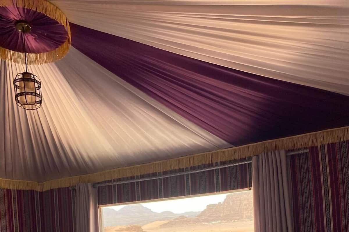 Una habitación con cama, dos sillas, una mesita y una ventana que da a unas tiendas de campaña vistas desde fuera.