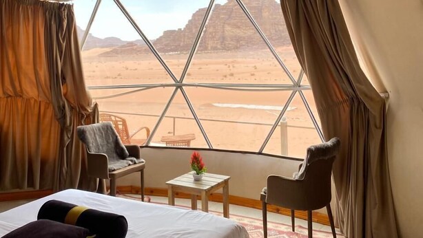 Una habitación con una cama, dos sillas, una mesita con un florero y vistas a un paisaje desértico a través de un ventanal grande.
