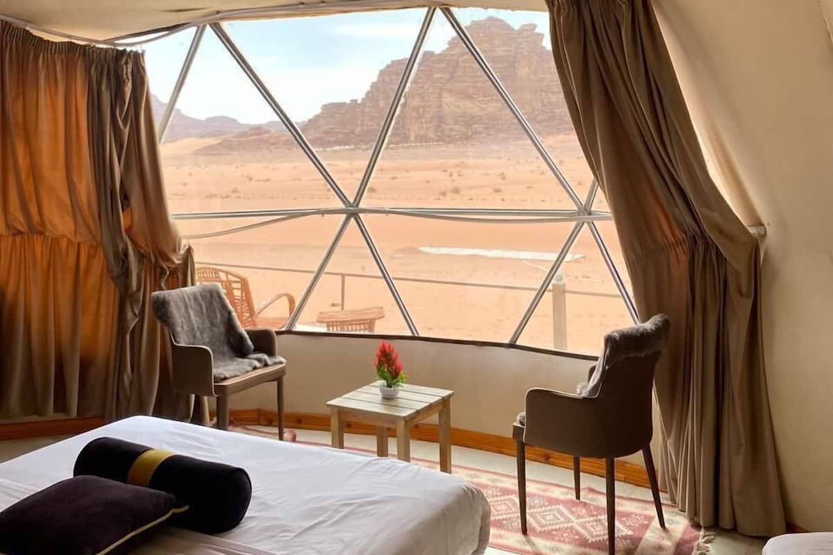 Una habitación con una cama, dos sillas, una mesita con un florero y vistas a un paisaje desértico a través de un ventanal grande.