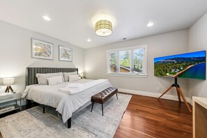 A bedroom with a bed, bedside tables, a bench, a TV, and a window with blinds.