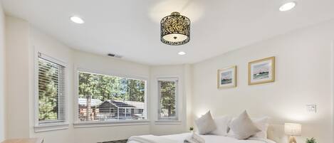A bedroom with a large bed, a bench, a bedside table with a lamp, and two windows with blinds.