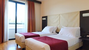 A hotel room with two beds, a large window with curtains, a nightstand, and a view of a balcony. - Raya Hotel (Mediglia)