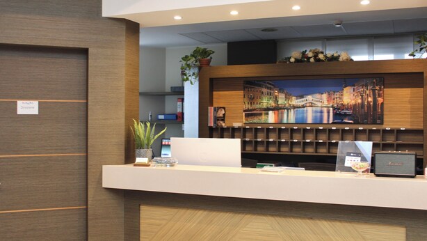 A reception desk with a modern design, a large framed picture of a cityscape, and a potted plant. - Raya Hotel (Mediglia)