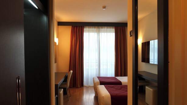 Habitación de hotel con cama, escritorio, silla, televisor y ventana con cortinas.