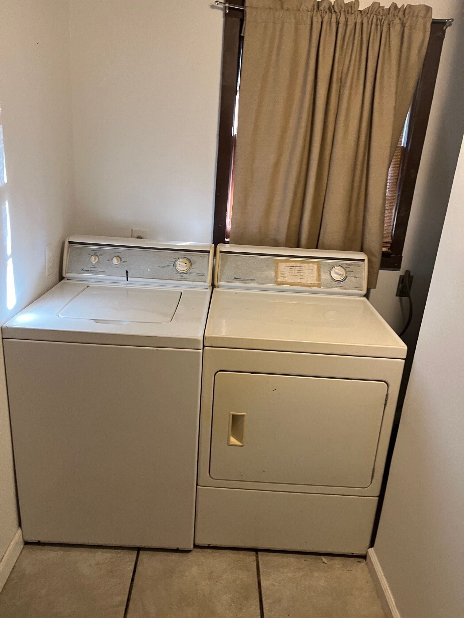 Un appareil compact combinant lave-linge et sèche-linge, à deux portes, installé dans une petite buanderie dotée d’une fenêtre et de rideaux.