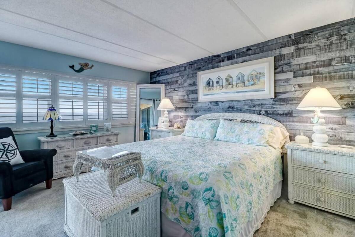 Une chambre à coucher avec un grand lit, deux tables de chevet, une chaise, un panier en osier et une fenêtre avec des stores.