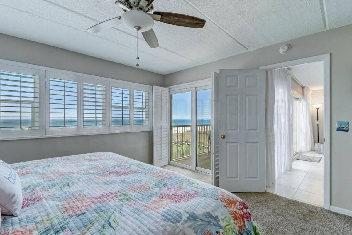 Une chambre avec un lit, un ventilateur de plafond, un balcon donnant sur l’océan et une porte menant à une autre pièce.