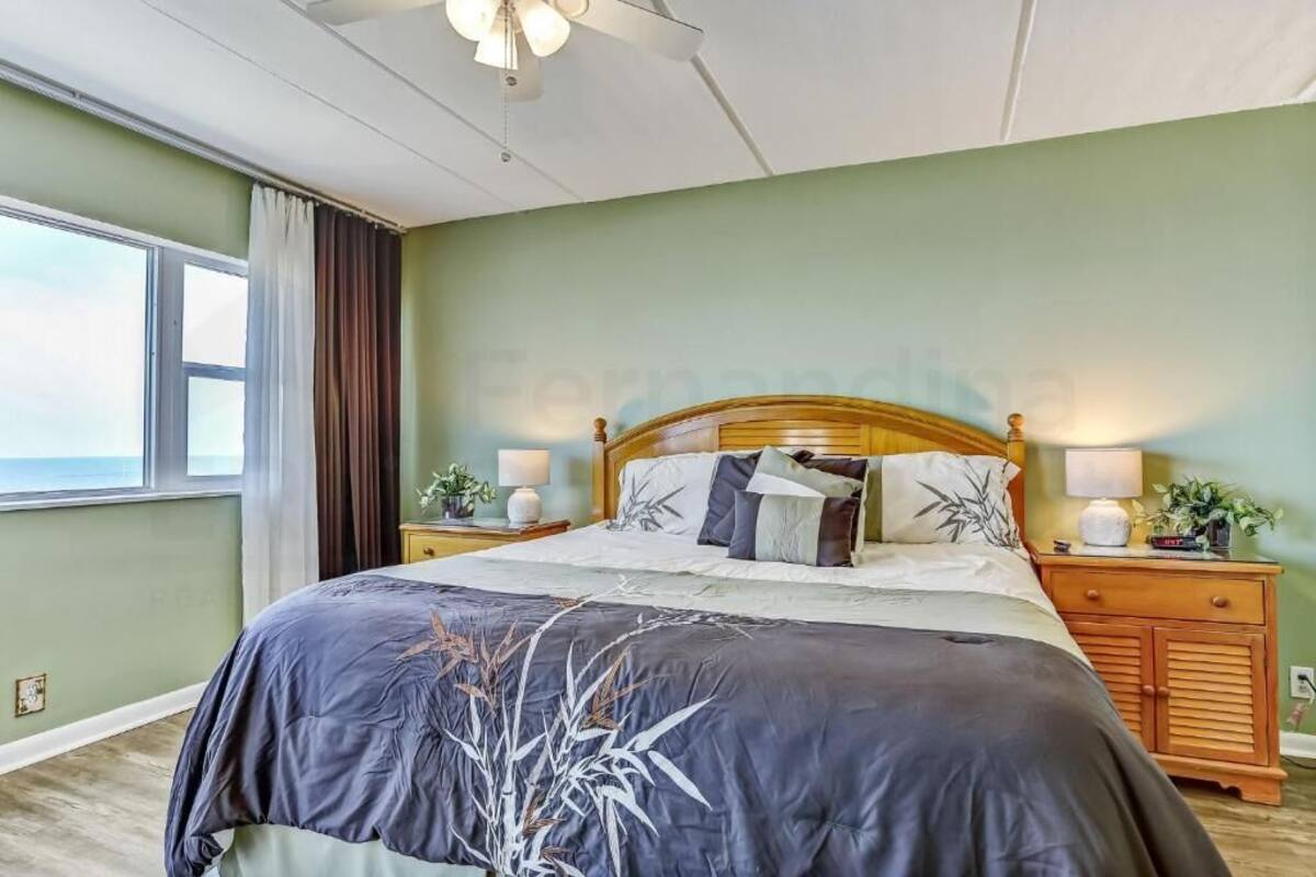 Une chambre à coucher avec un grand lit, deux tables de chevet avec des lampes, une tête de lit en bois et une vue sur l’extérieur.