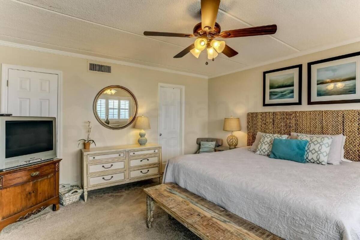 Une chambre à coucher comprenant un lit, un ventilateur de plafond, une commode, un téléviseur, un miroir et deux tableaux encadrés accrochés au mur.