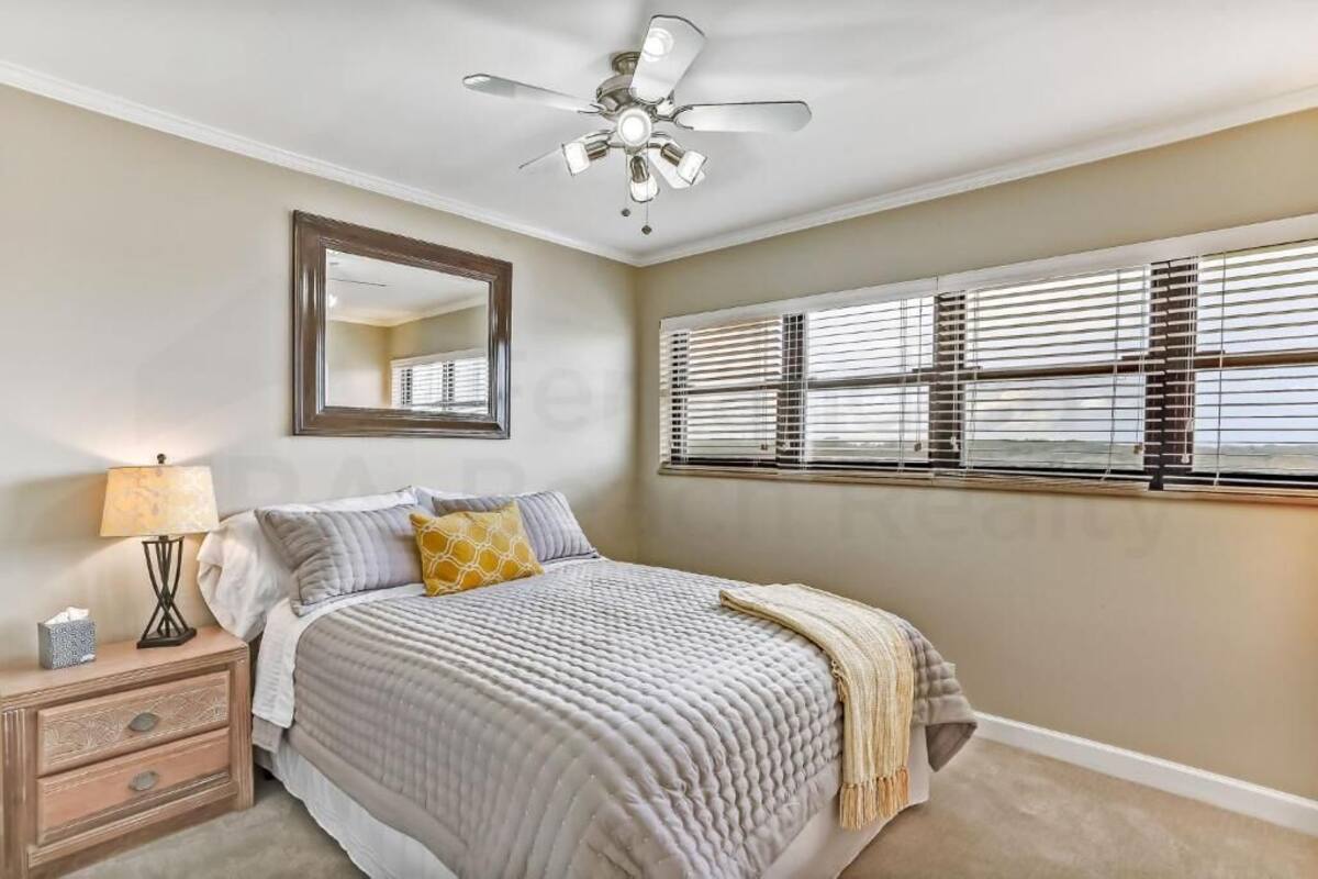 Une chambre à coucher comprenant un lit, une table de chevet, une lampe, un ventilateur de plafond et une fenêtre avec des stores.