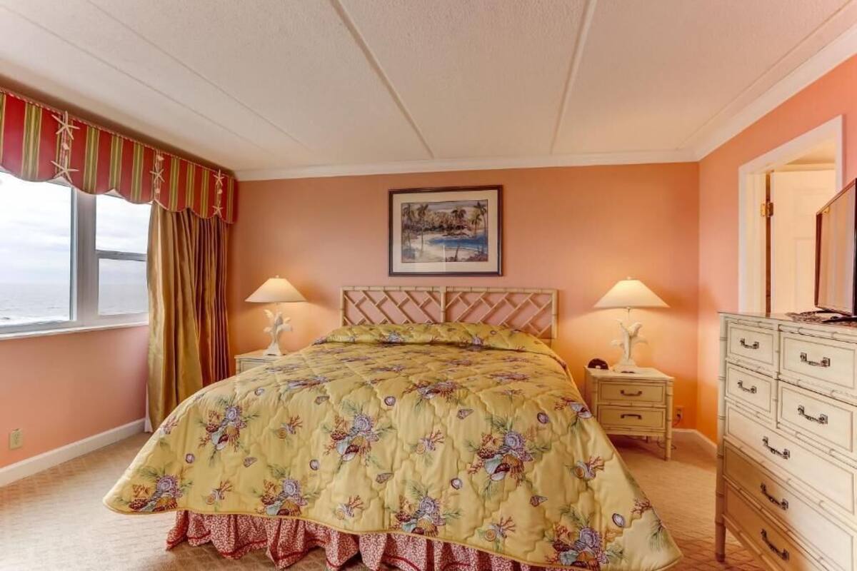 Une chambre à coucher avec un lit, deux tables de chevet avec des lampes, une commode, une fenêtre avec des rideaux et un tableau au mur.