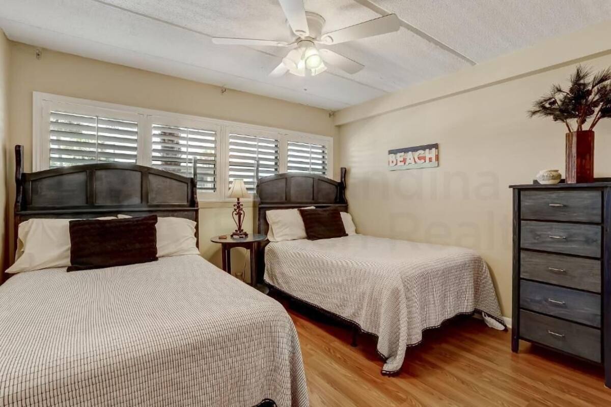 Une chambre avec deux lits, un ventilateur de plafond, une table de chevet, une commode et une fenêtre avec des stores.