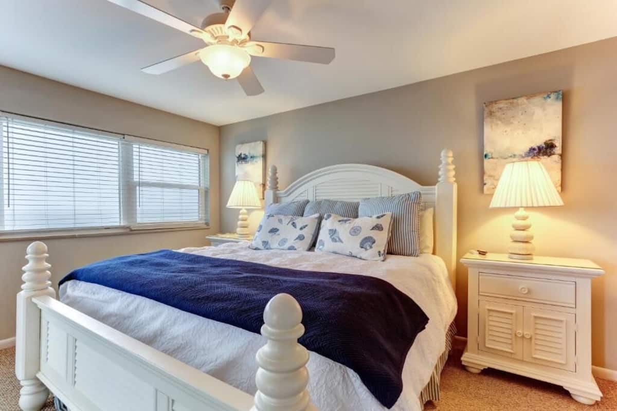 Une chambre à coucher avec un lit blanc, un ventilateur de plafond, une table de chevet avec une lampe et une fenêtre avec des stores.