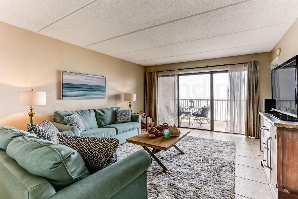 A living room with a green sofa, a coffee table, and a TV. There is a balcony with a view.