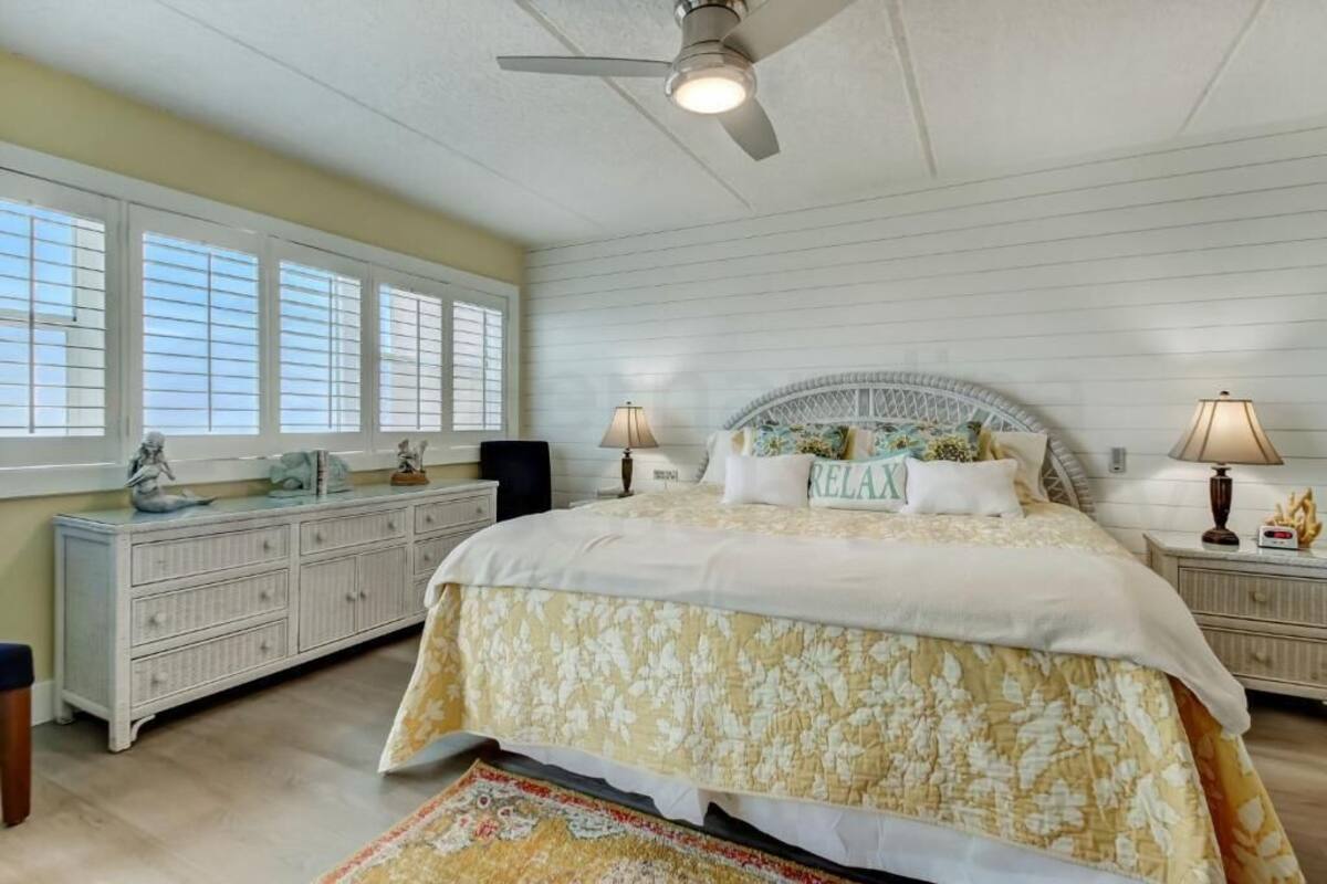 Une chambre à coucher comprenant un lit, deux tables de chevet, un ventilateur de plafond, une fenêtre avec stores et une tête de lit décorative.
