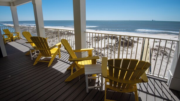 A wooden deck with yellow Adirondack chairs and a white table, overlooking a sandy beach and the ocean.
