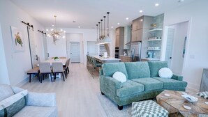 A modern kitchen with a central island, a dining area with a long table and chairs, and a living area with a sofa and ottoman.