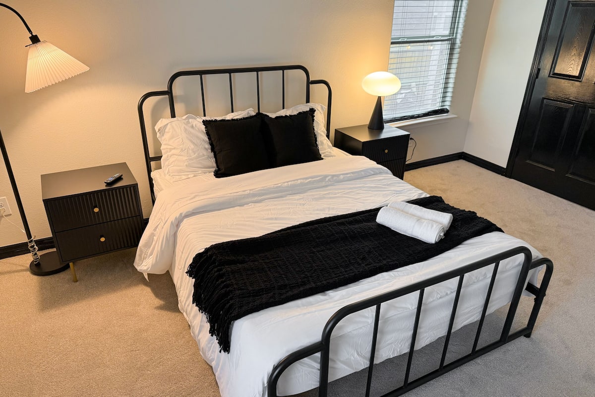 A neatly made bed with white and black bedding, a black throw blanket, and a rolled towel. There is a bedside lamp and a small bedside table with a remote control.