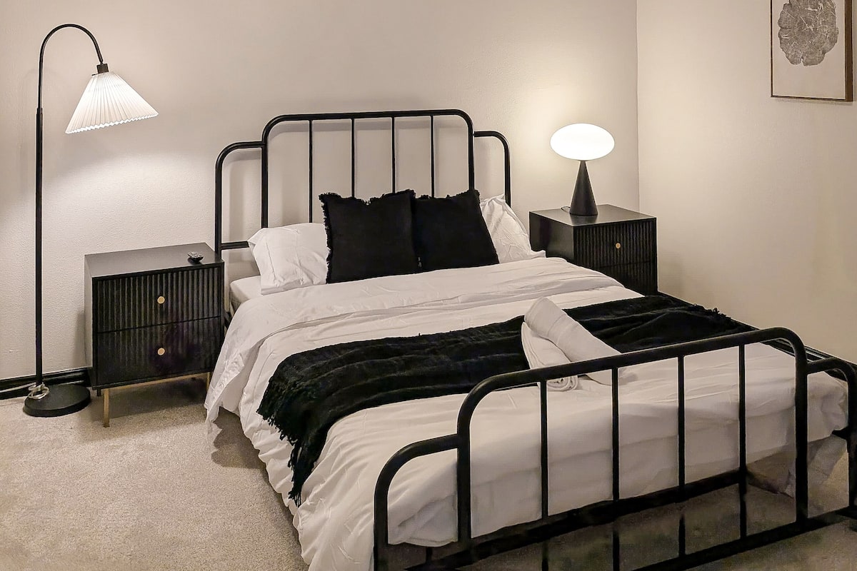 A bedroom with a metal bed frame, white bedding, a black throw blanket, a bedside table with a lamp, and a ceiling light fixture.