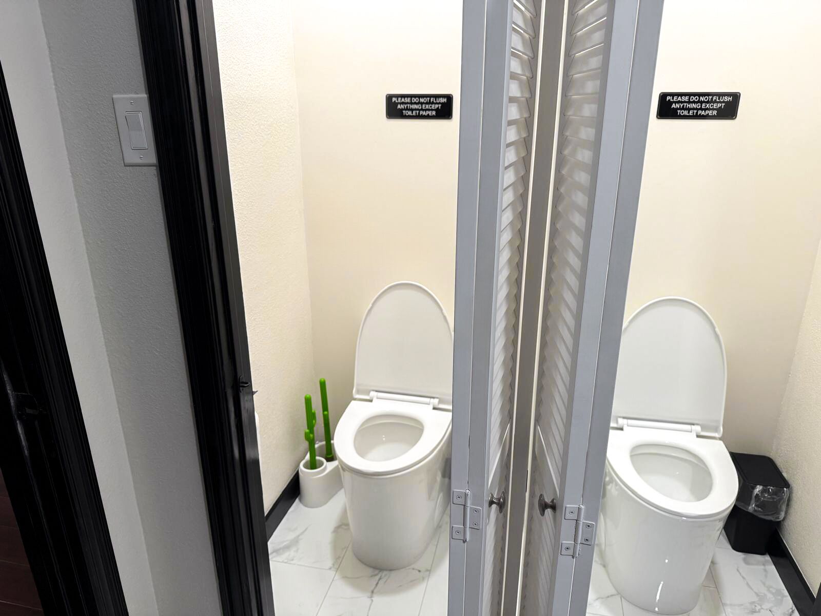 A modern bathroom with two toilets, each with a privacy partition and a sign instructing not to flush anything except toilet paper.