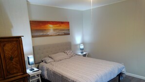 A bedroom with a bed, two bedside tables, a nightstand, a wooden dresser, and a painting of a sunset over the ocean.