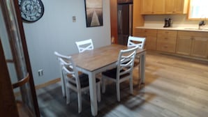 A dining area with a wooden table and white chairs, a refrigerator, and wooden cabinets in the background.