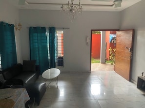 Uma sala de estar com sofá, mesa redonda, lustre e vista para um jardim por uma porta aberta.