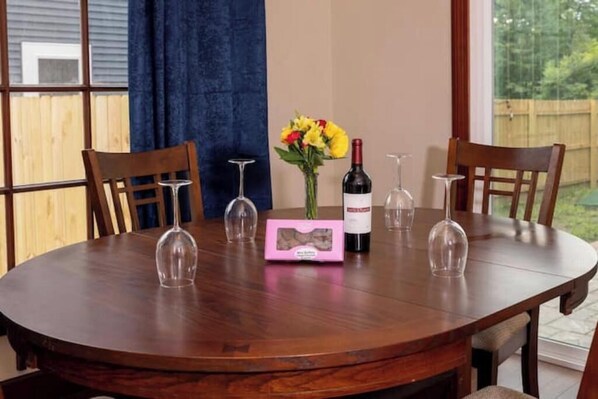 A round wooden dining table with four chairs, a vase with yellow flowers, a wine bottle, and a pink box with a picture.