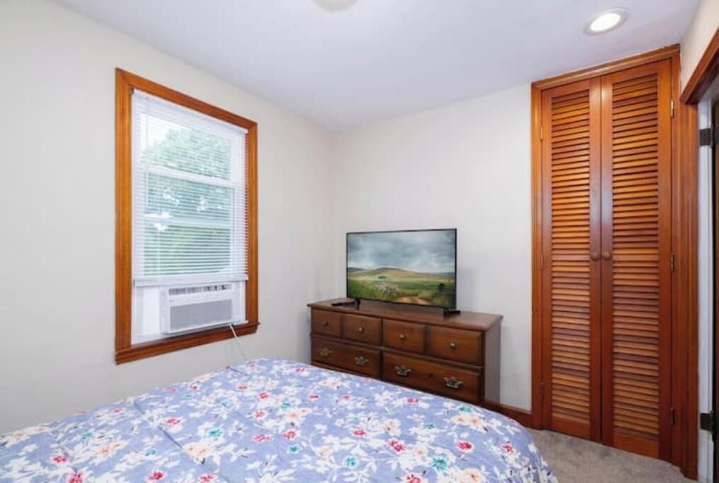 Un dormitorio con cama, cómoda, televisor, ventana con persianas y armario de madera.