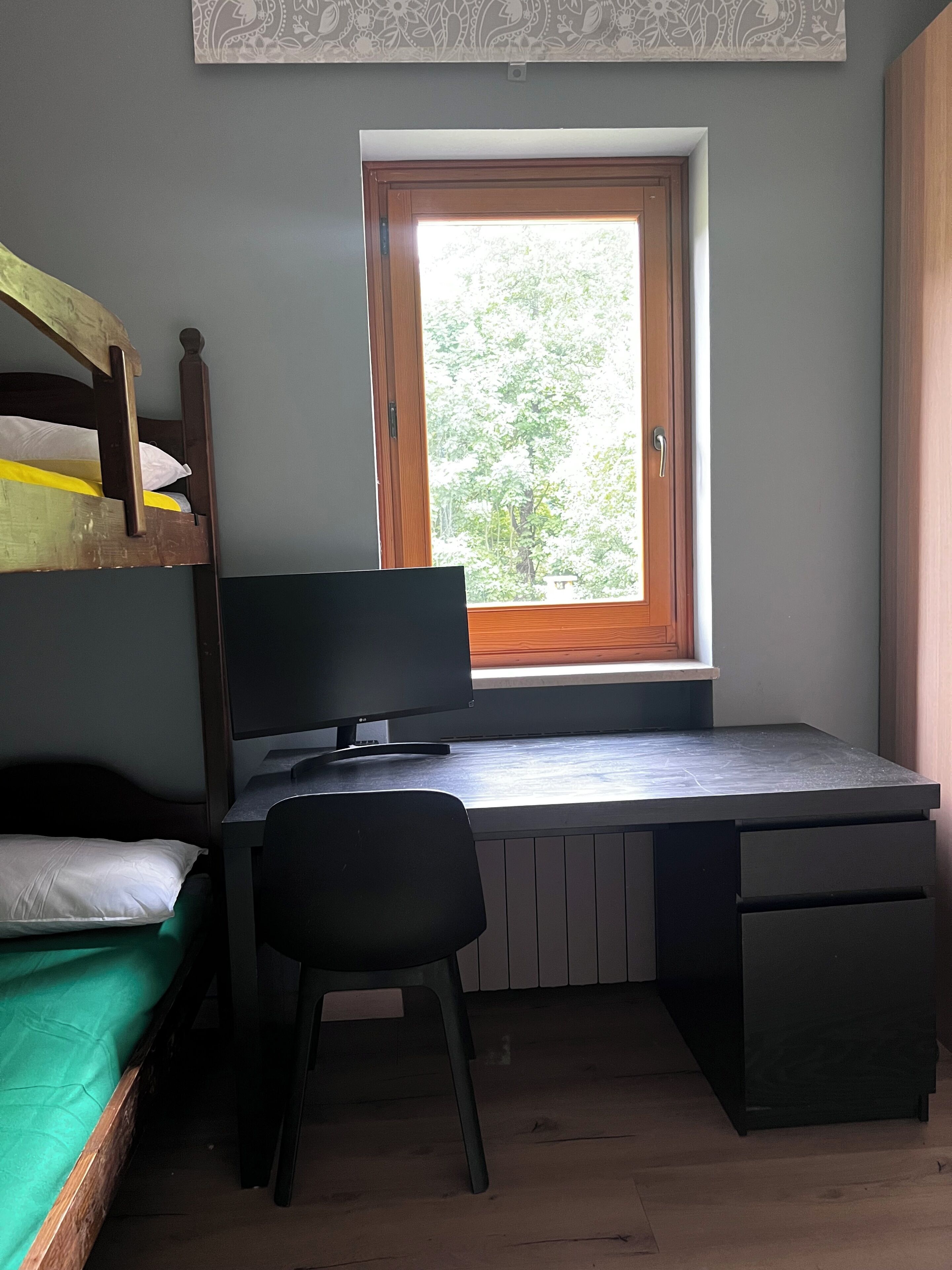 Ein Schlafraum im Studentenwohnheimstil mit Etagenbett, Schreibtisch inklusive Computer und Fenster mit Blick auf Bäume.