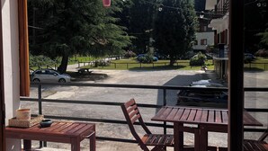 Een balkon met een houten tafel en stoelen, een witte bank en uitzicht op een parkeerplaats met auto's en bomen.