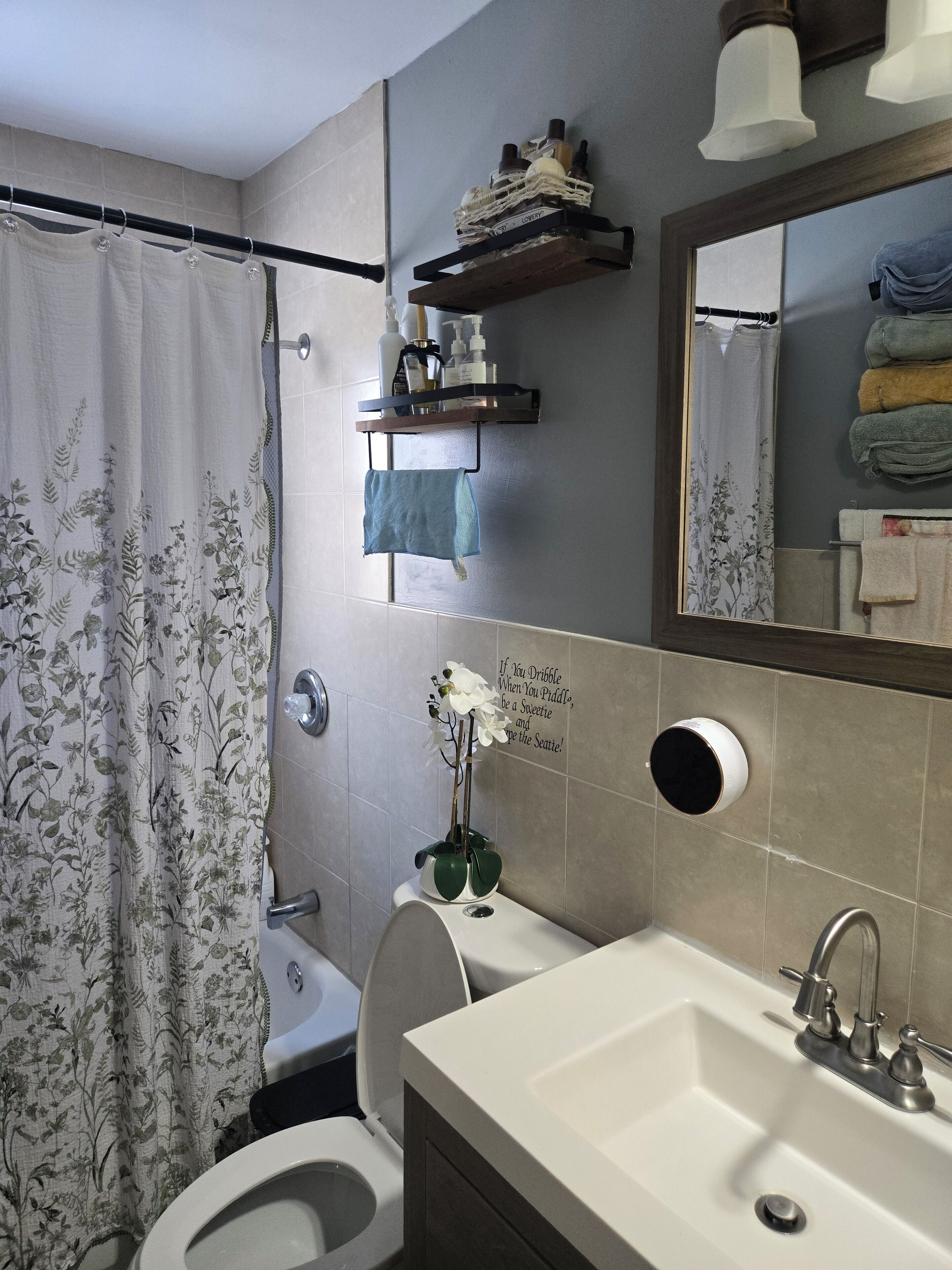 A bathroom with a toilet, sink, and shower curtain. There is a shelf with toiletries and a mirror above the sink.